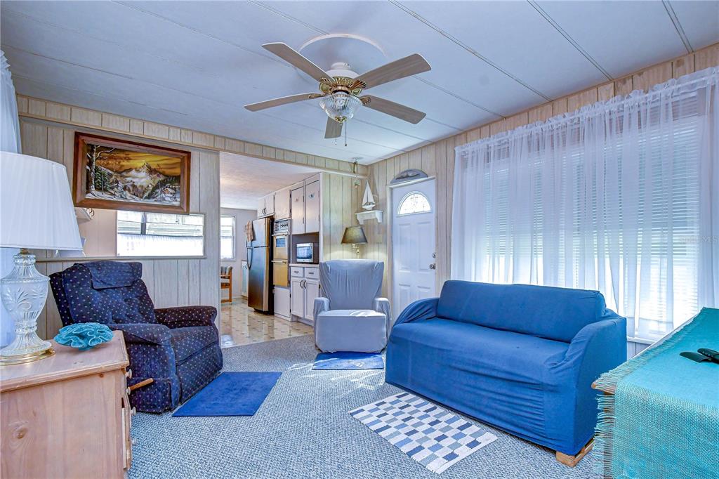 A living room features blue furniture, wood-paneled walls, a ceiling fan, and a kitchen view — 38651 WINDFLOWER AVENUE, ZEPHYRHILLS, FL
