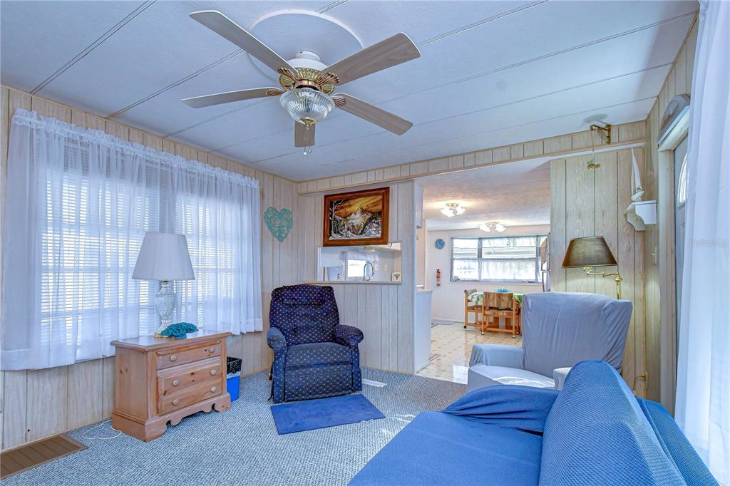 A living room features wood-paneled walls, a ceiling fan, blue furniture, and a carpeted floor — 38651 WINDFLOWER AVENUE, ZEPHYRHILLS, FL