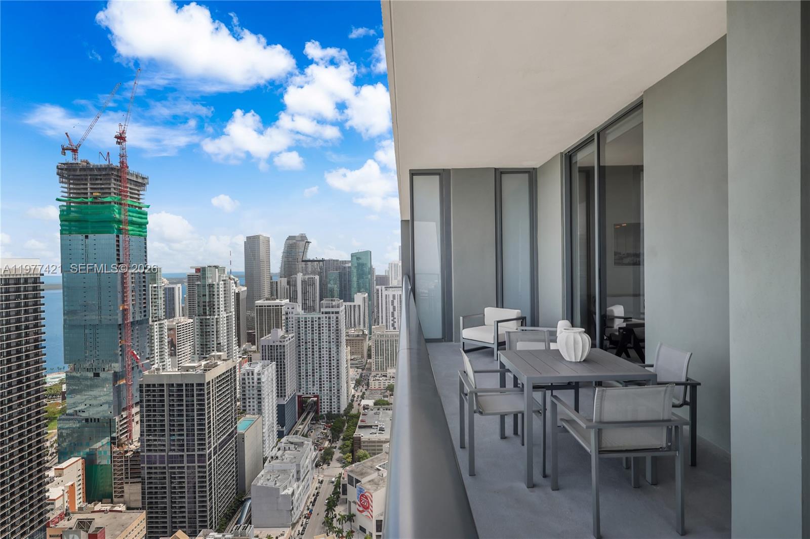 A high-rise balcony features outdoor furniture overlooking a dense city skyline with a construction crane — 159 NE 6th St # 5115, Miami FL 33132, MIAMI, FL