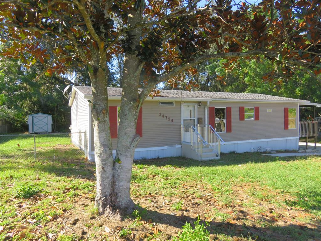 A single-story manufactured home with tan siding and red shutters sits behind a large tree — 14314 SE 58TH COURT, SUMMERFIELD, FL