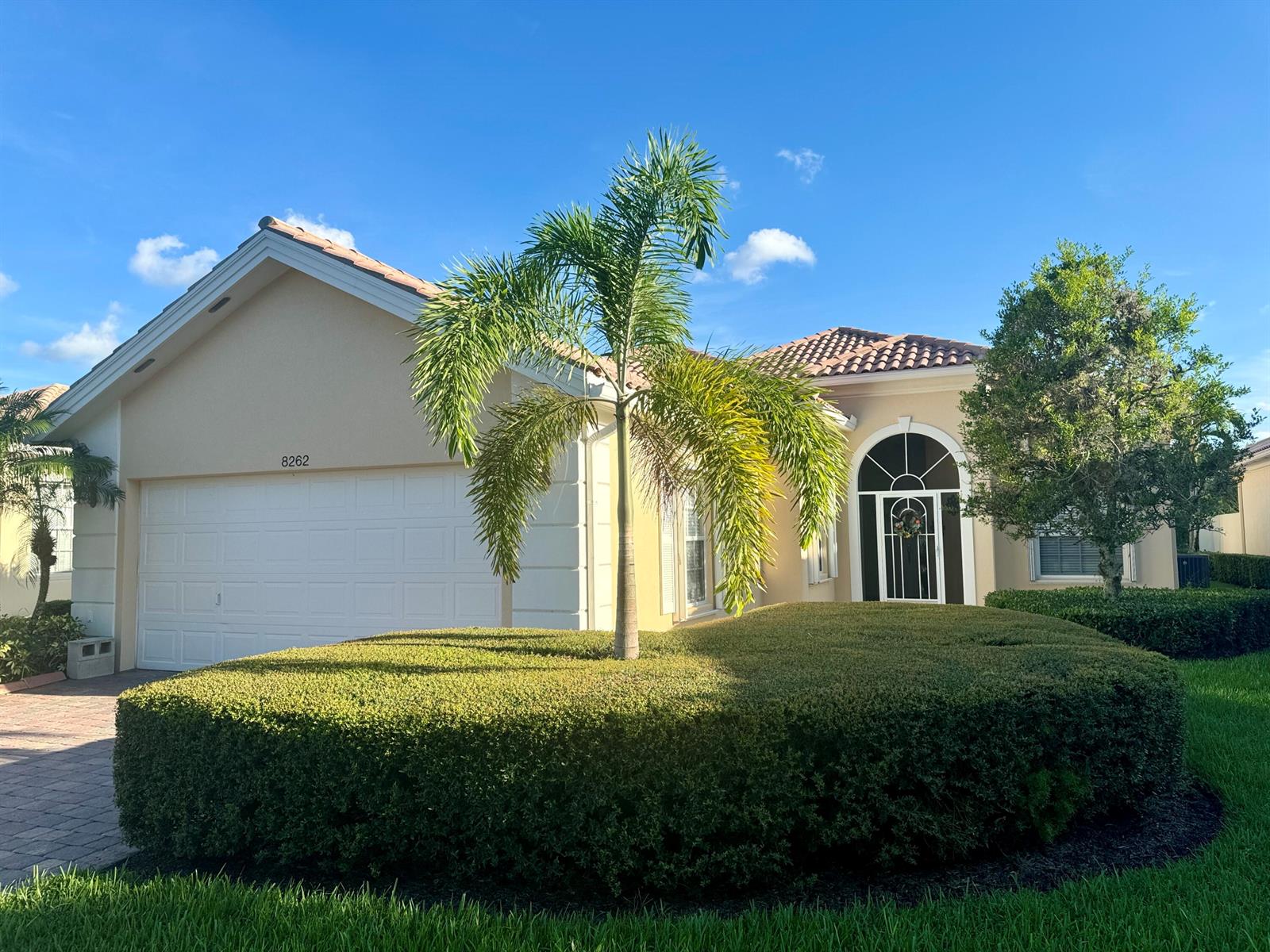 A single-story house with a beige stucco exterior, a palm tree, and a large hedge — 8262 Dominica Place, Wellington FL 33414, WELLINGTON, FL