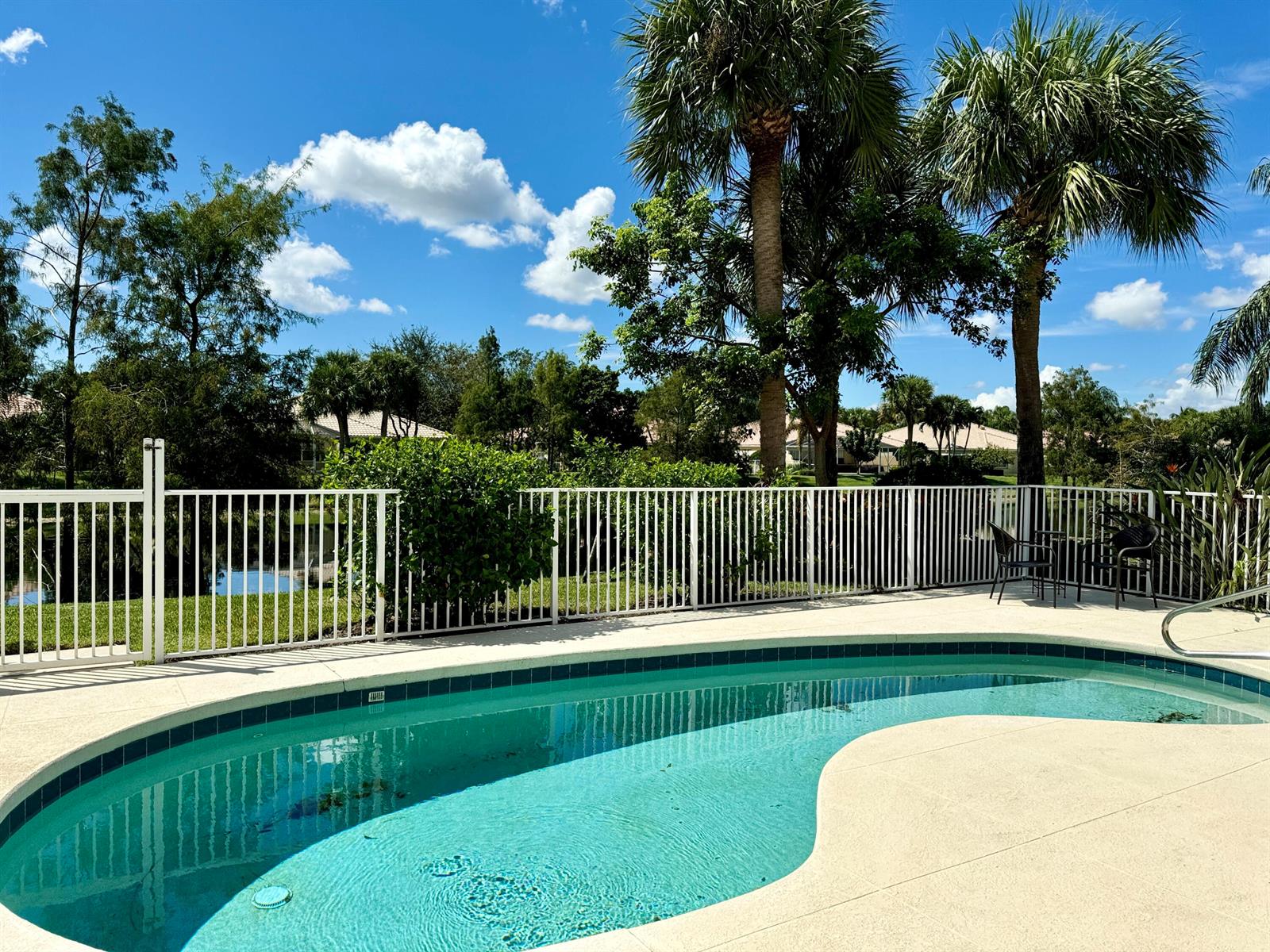 A kidney-shaped swimming pool sits on a patio behind a white fence and palm trees — 8262 Dominica Place, Wellington FL 33414, WELLINGTON, FL