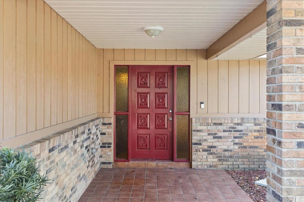 A red front door with side windows sits under a porch with brick and siding — 2047 ROCKY HILL DRIVE, DELTONA, FL