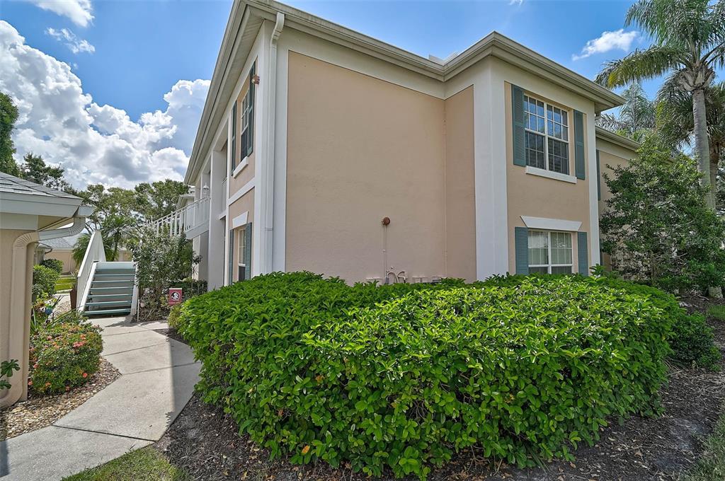 A two-story apartment building exterior with beige stucco walls, green shutters, and a large hedge — 4705 SAND TRAP STREET CIRCLE E UNIT 106, BRADENTON, FL