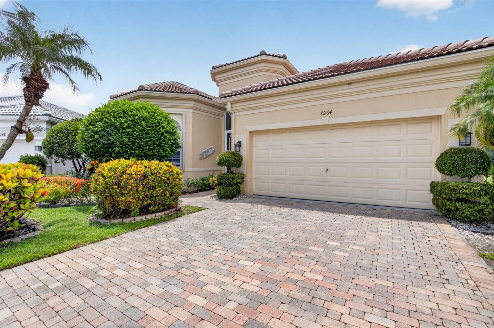 The exterior of a tan house features a paver driveway, a two-car garage, and landscaping — 7264 Cataluna Circle, Delray Beach FL 33446, DELRAY BEACH, FL