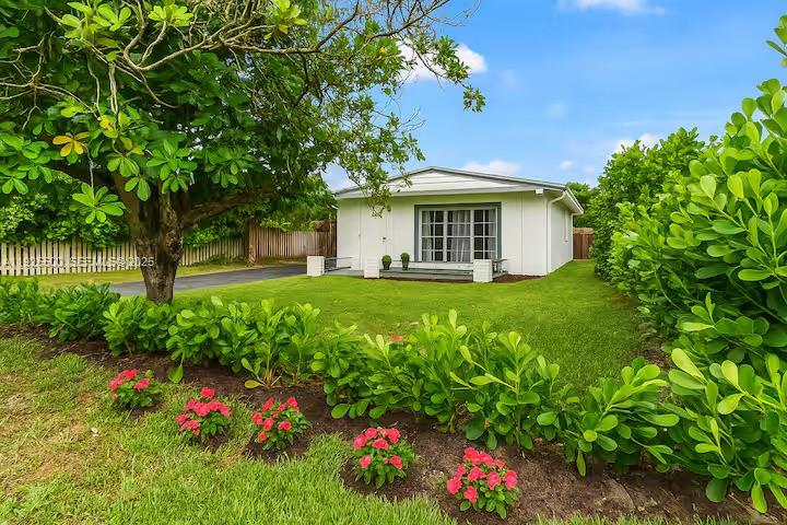 A white single-story house sits behind a green lawn with a large tree and flowers — 909 NE 7th St, Pompano Beach FL 33060, POMPANO BEACH, FL