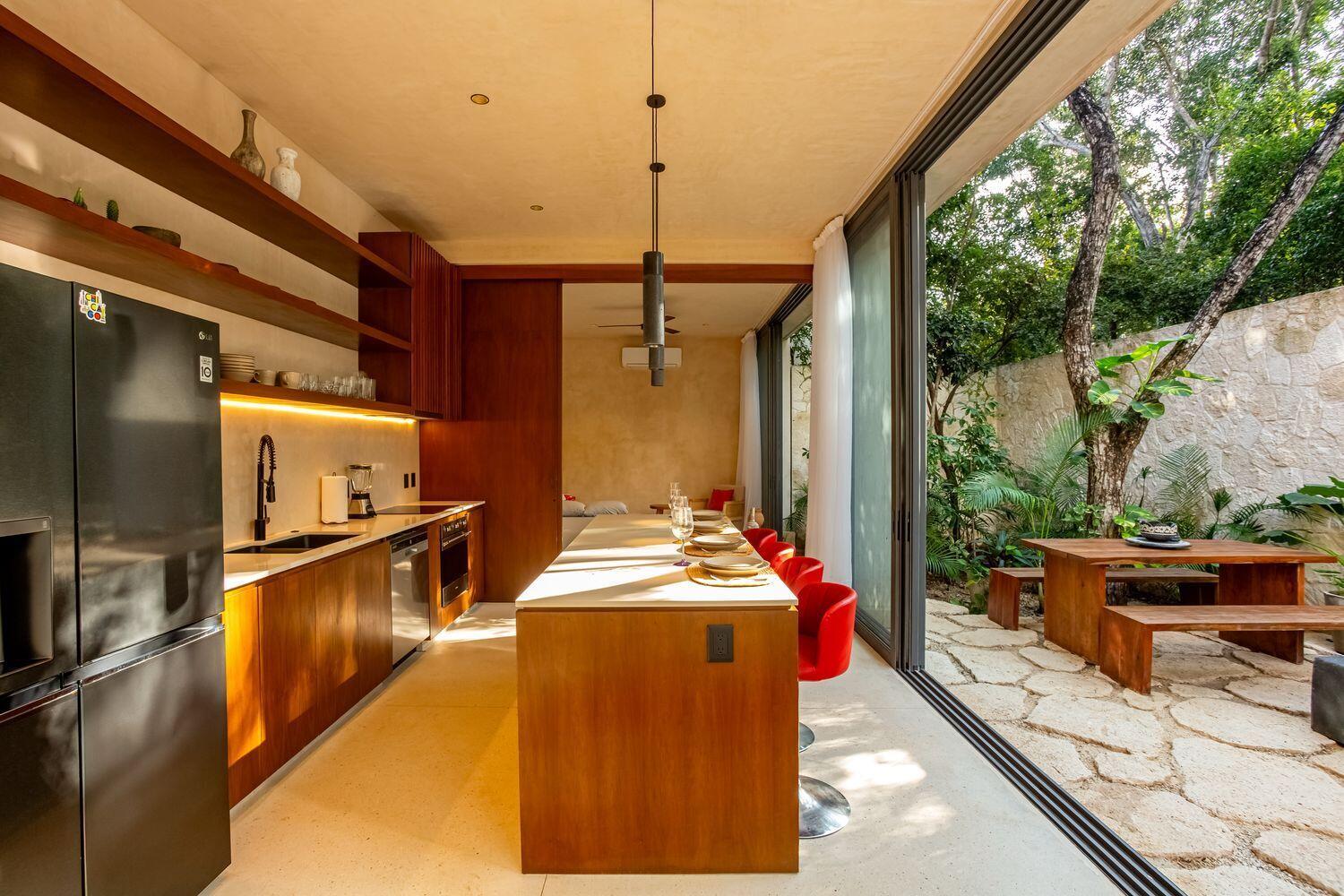 A modern kitchen with wood cabinetry and an island opens to an outdoor stone patio — Amari Tulum 10 Avenida Norte & Calle Palma, FL