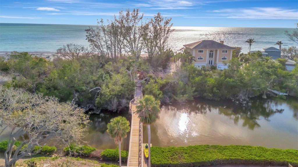 An aerial view shows a wooden footbridge crossing a pond toward a house near the ocean — 8555 HERON LAGOON CIRCLE, SARASOTA, FL