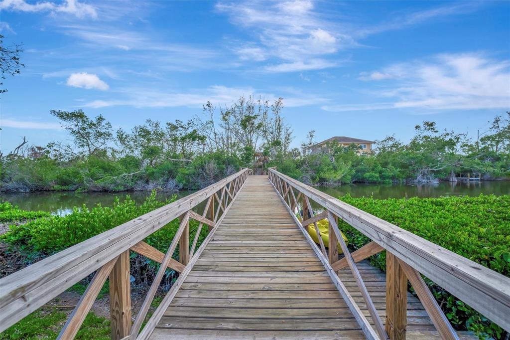 A wooden footbridge with railings extends over a narrow waterway surrounded by lush green vegetation — 8555 HERON LAGOON CIRCLE, SARASOTA, FL