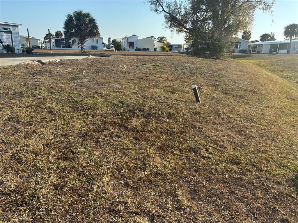A vacant, grassy residential lot features a utility pipe and surrounding mobile homes — 7437 LONGMONT LANE, PORT CHARLOTTE, FL