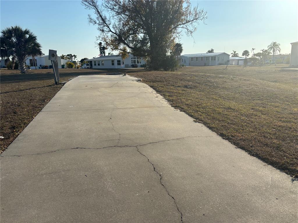 A cracked concrete driveway leads toward a vacant lot with a large tree and houses — 7437 LONGMONT LANE, PORT CHARLOTTE, FL