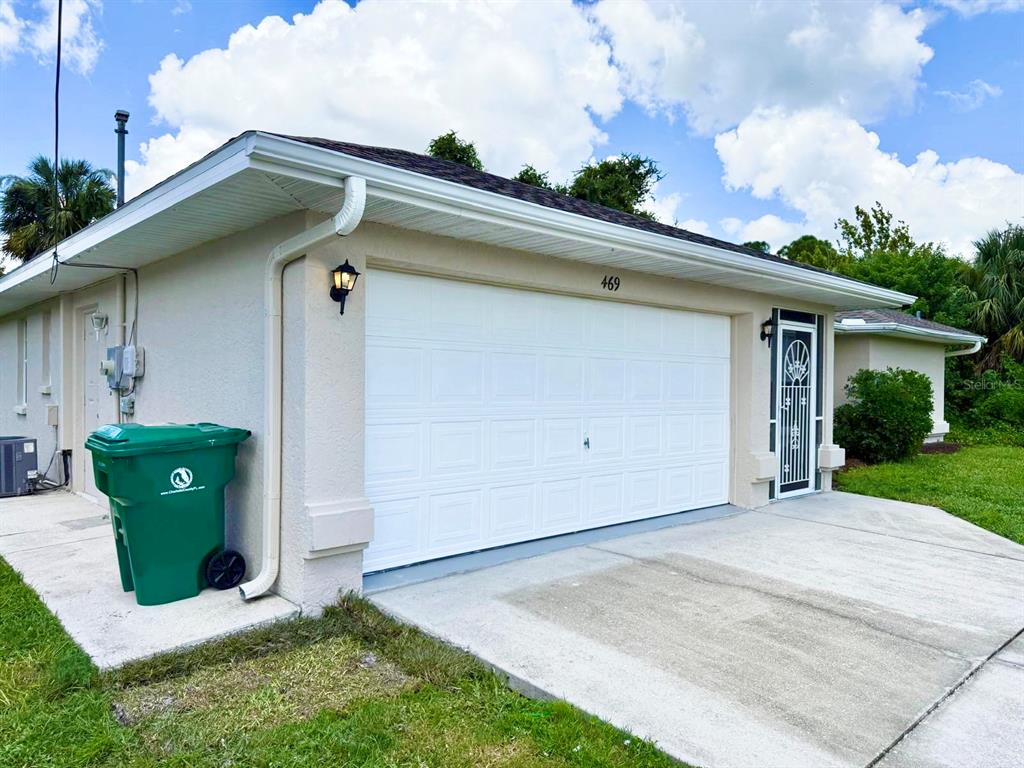 A beige house exterior features a white garage door, a green trash bin, and landscaping — 469 DORCHESTER STREET, PORT CHARLOTTE, FL