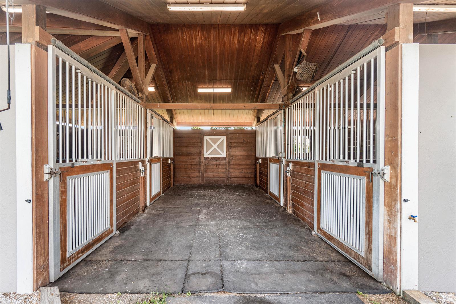 A stable aisle features wooden stalls with white metal bars and rubber mat flooring — 2080 Canter Way, Wellington FL 33414, WELLINGTON, FL