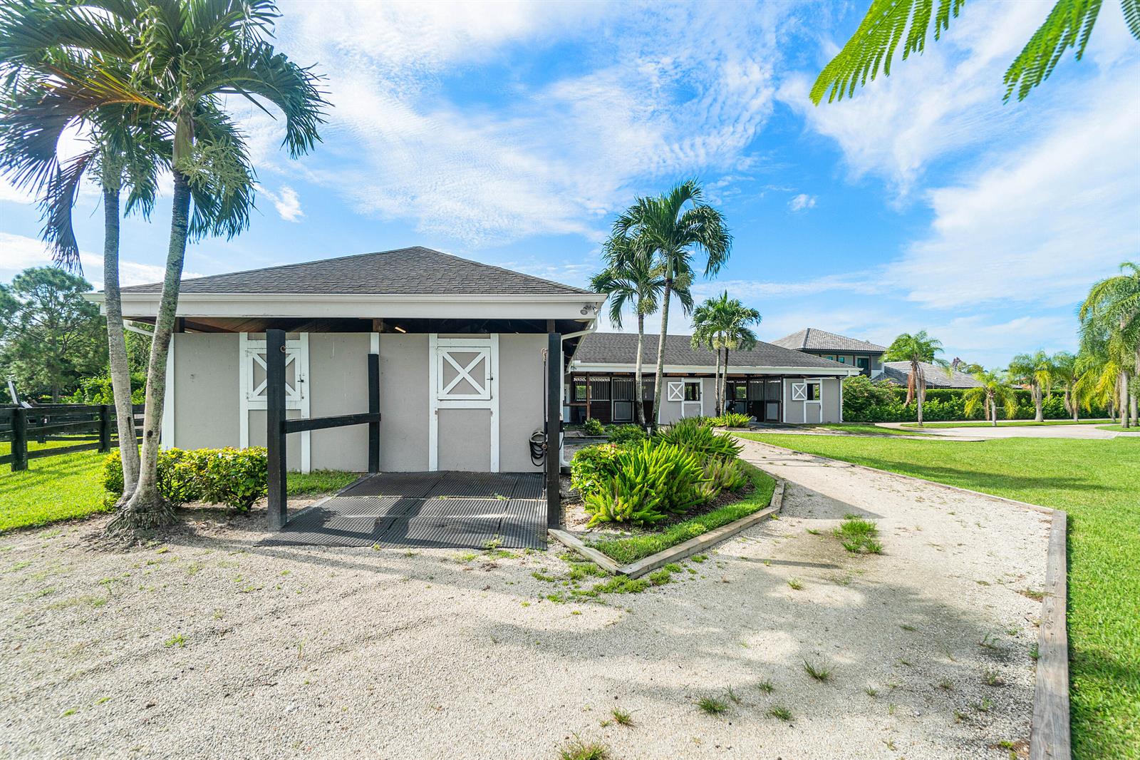 A gravel driveway leads to a gray horse stable with white doors and palm trees — 2080 Canter Way, Wellington FL 33414, WELLINGTON, FL