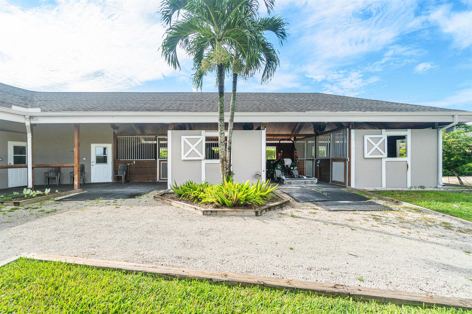 An exterior view of a horse stable with stalls, a palm tree, and gravel ground — 2080 Canter Way, Wellington FL 33414, WELLINGTON, FL