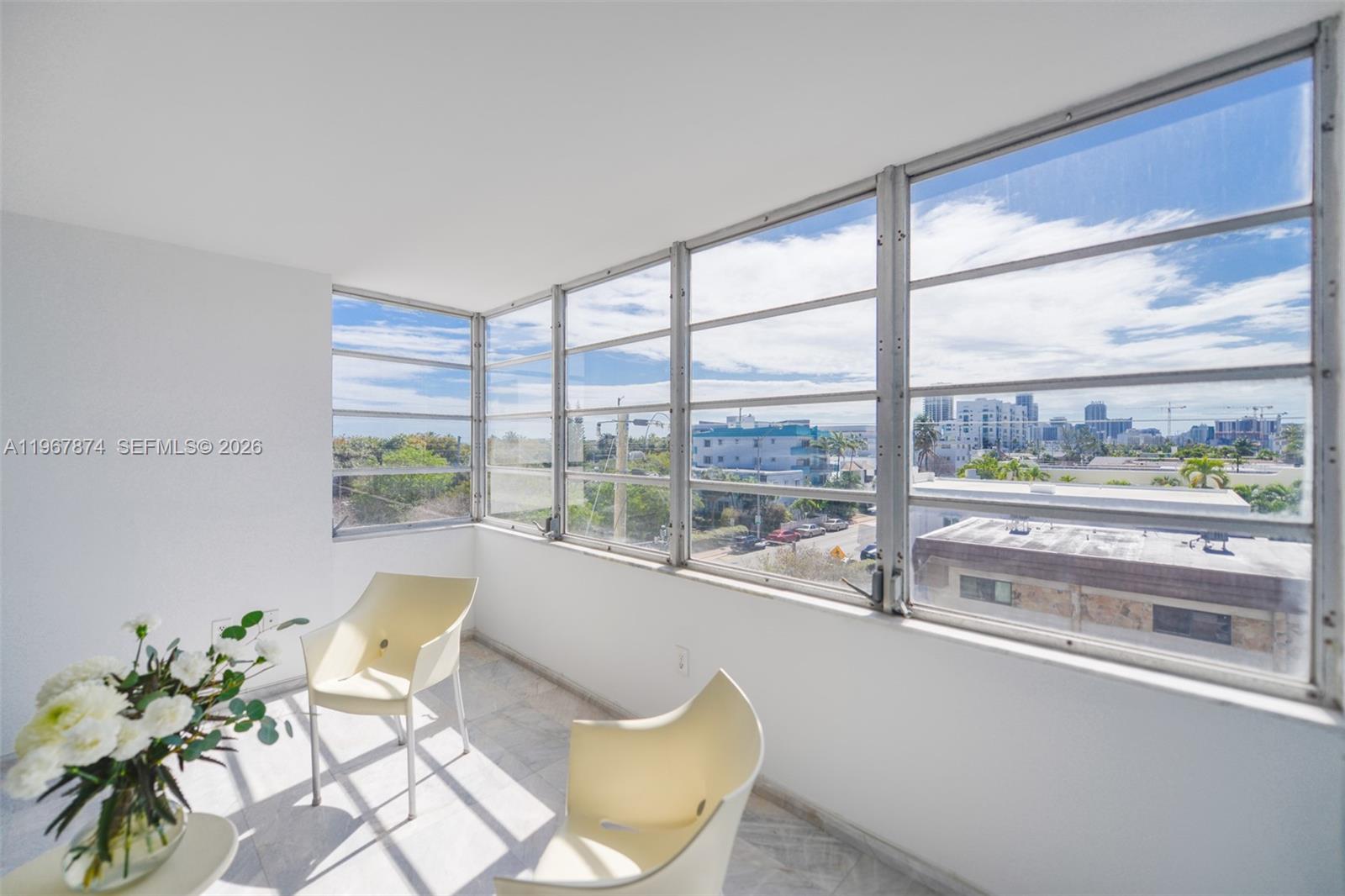 A sunroom features white walls, marble flooring, two chairs, and large windows overlooking a city — 8000 Harding Ave # 4B, Miami Beach FL 33141, MIAMI BEACH, FL