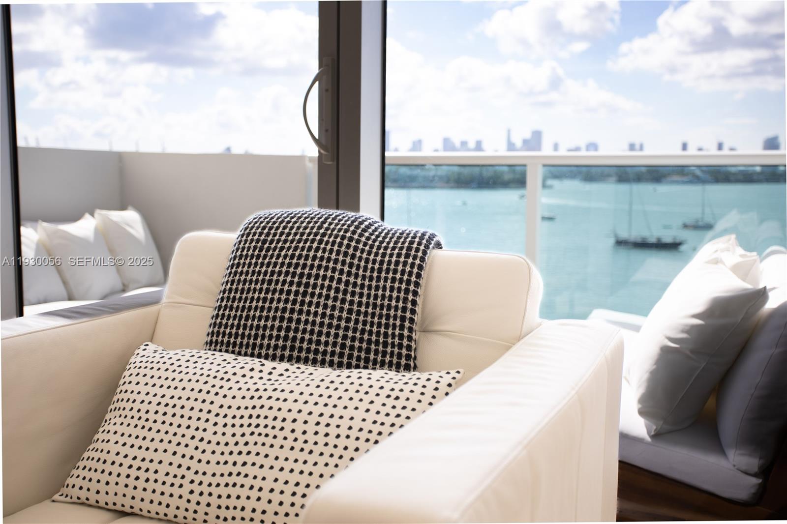 A white armchair with patterned pillows sits before a balcony overlooking a bay and skyline — 1200 West Ave # 526, Miami Beach FL 33139, MIAMI BEACH, FL