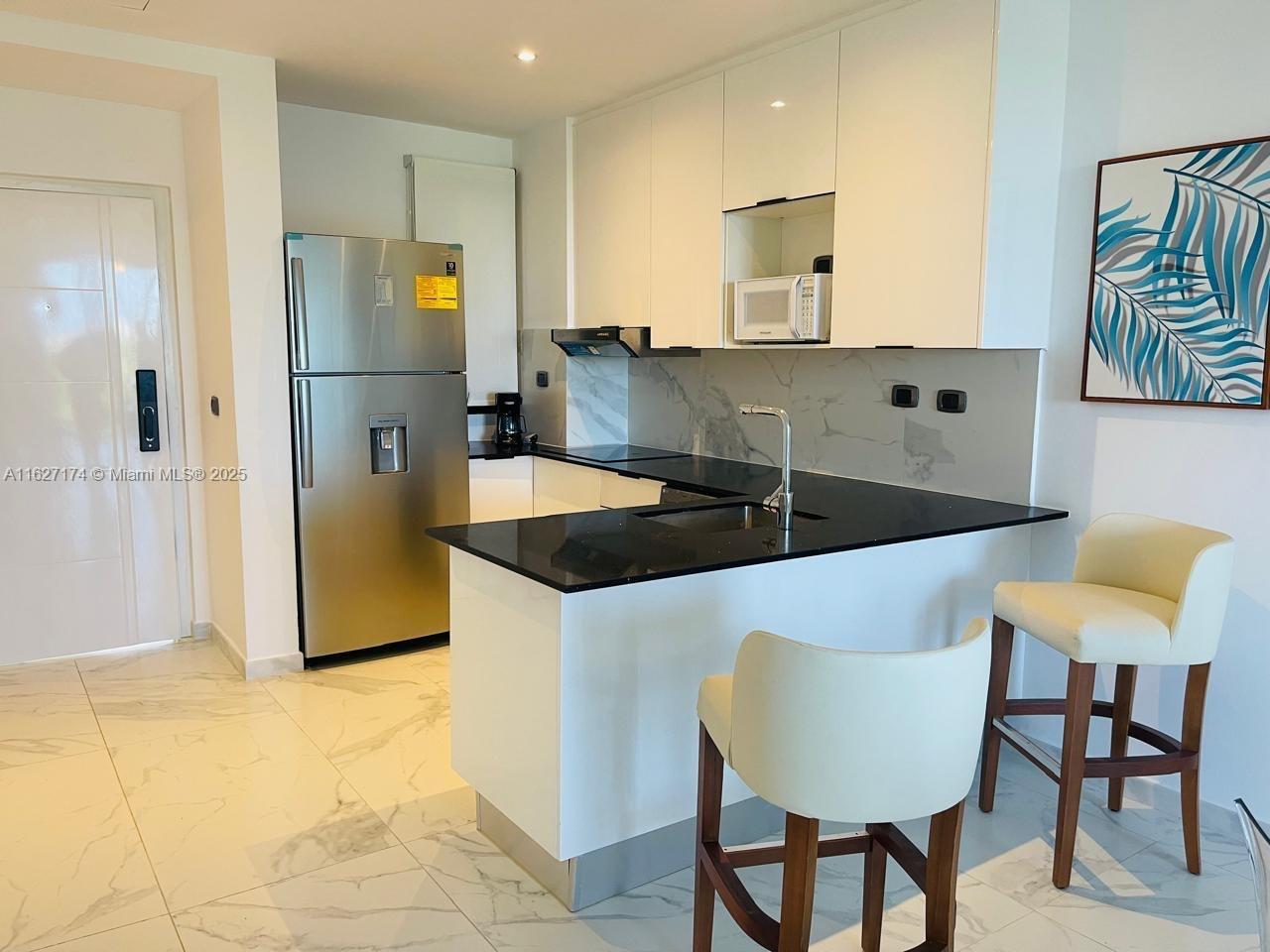 A modern kitchen features white cabinets, black countertops, stainless steel appliances, and two bar stools — Av. Hard Rock Punta Cana 23000 #PH-510 , Dominican Republic, DR, DR