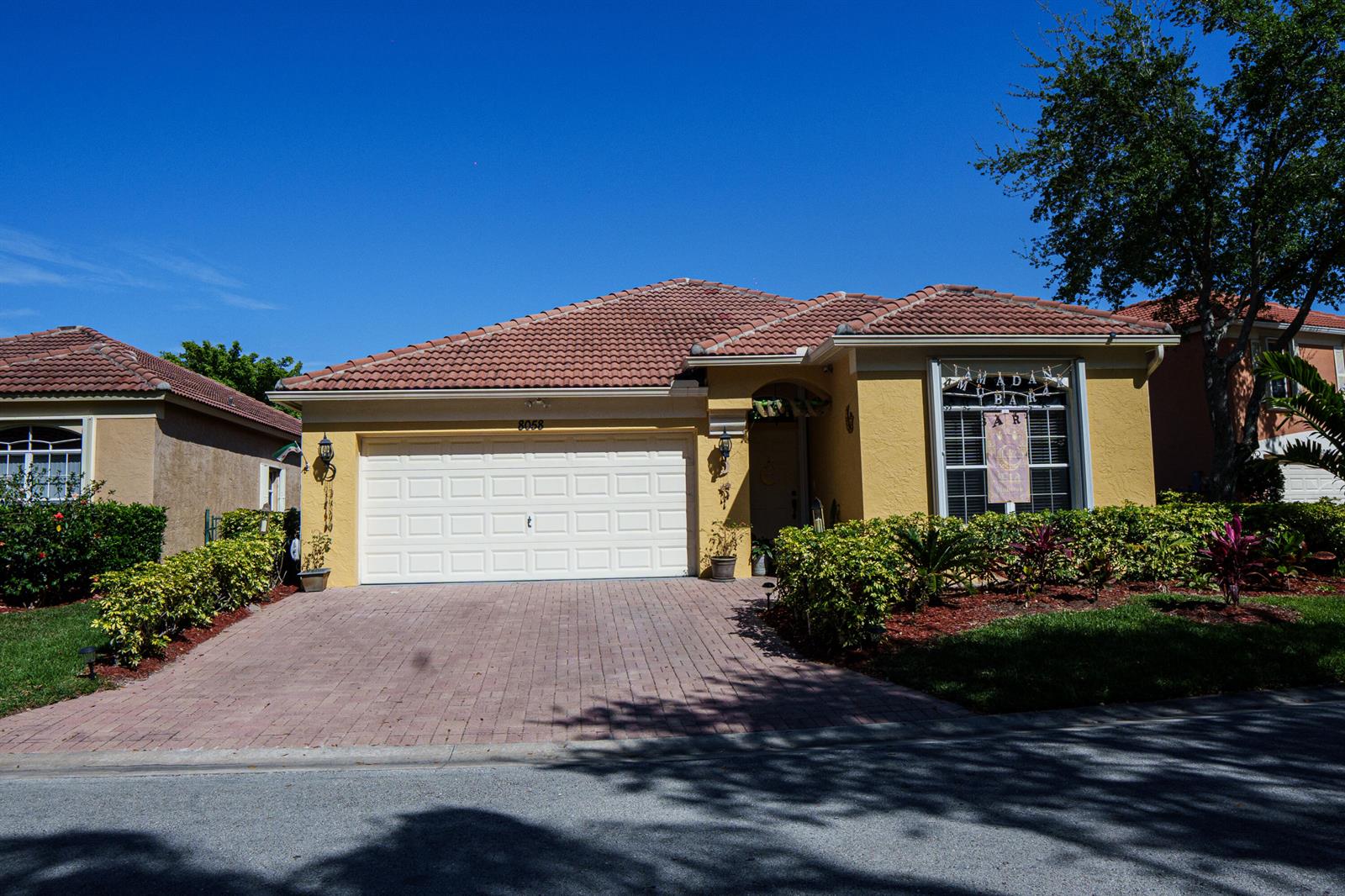 A single-story yellow stucco house with a tiled roof, a two-car garage, and landscaped front yard — 8058 Via Hacienda, Riviera Beach FL 33418, RIVIERA BEACH, FL