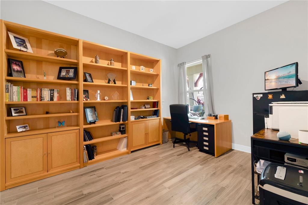 A home office features light wood built-in shelving, a desk, and wood-look tile flooring — 16429 FREHLEY RUN, LAND O LAKES, FL