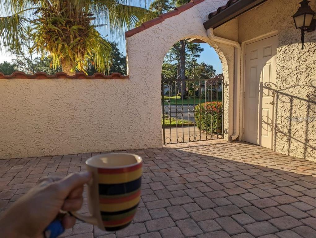 A hand holds a striped mug on a paved patio with a gated arched entryway — 5581 GOLF POINTE DRIVE, SARASOTA, FL