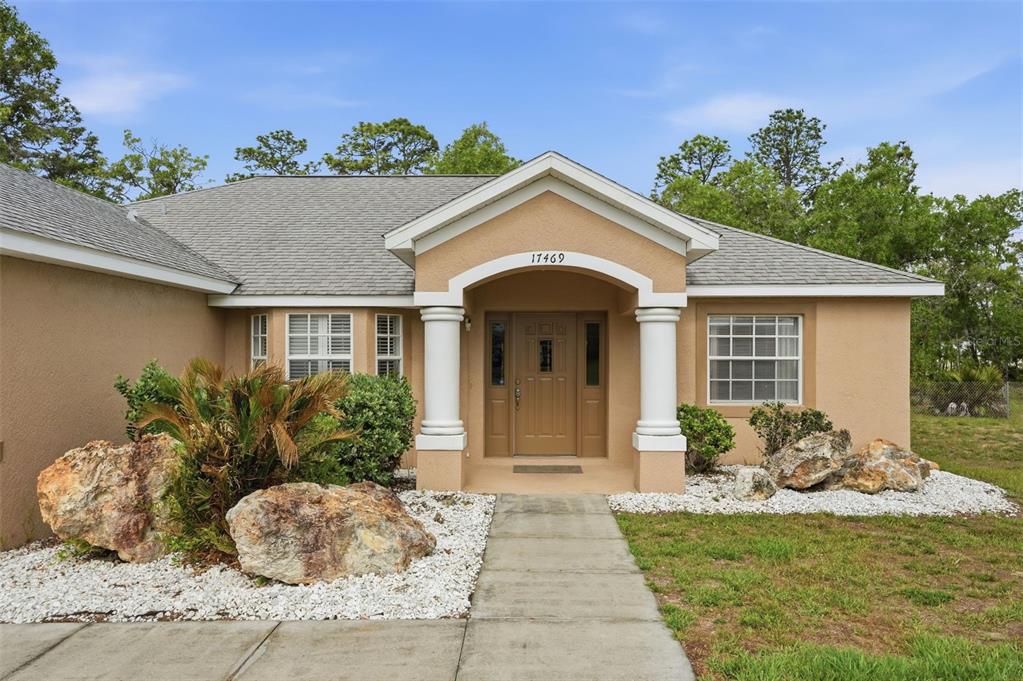 A tan house exterior features a covered entryway with white columns and a concrete walkway — 17469 MABERLY ROAD, WEEKI WACHEE, FL