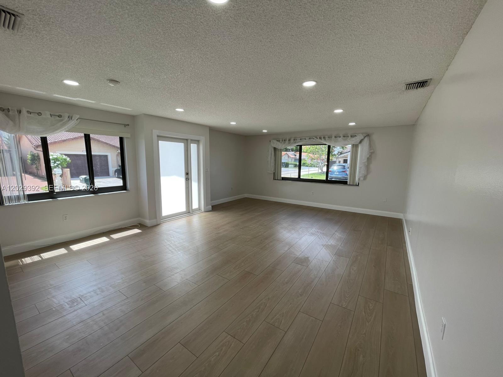 An empty living room features light wood-look flooring, recessed lighting, and two large windows — 1727 SW 136th Pl, Miami FL 33175, MIAMI, FL