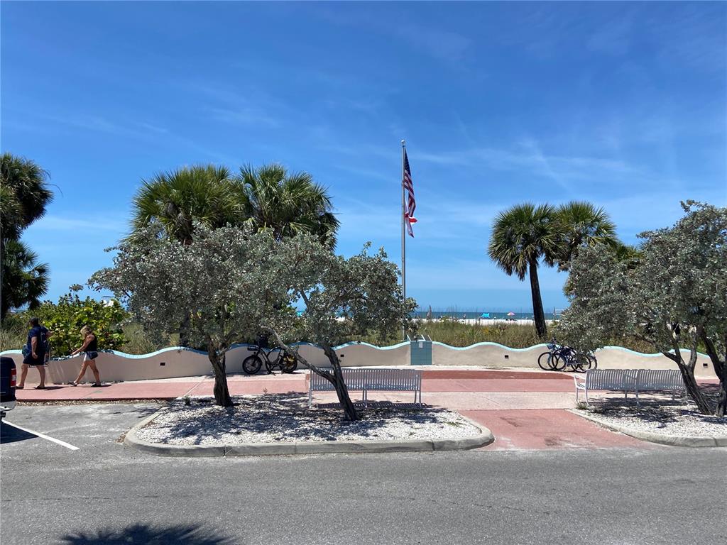 A paved path with benches and palm trees leads toward a beach with an American flag — 605 69TH AVENUE UNIT 1, ST PETE BEACH, FL