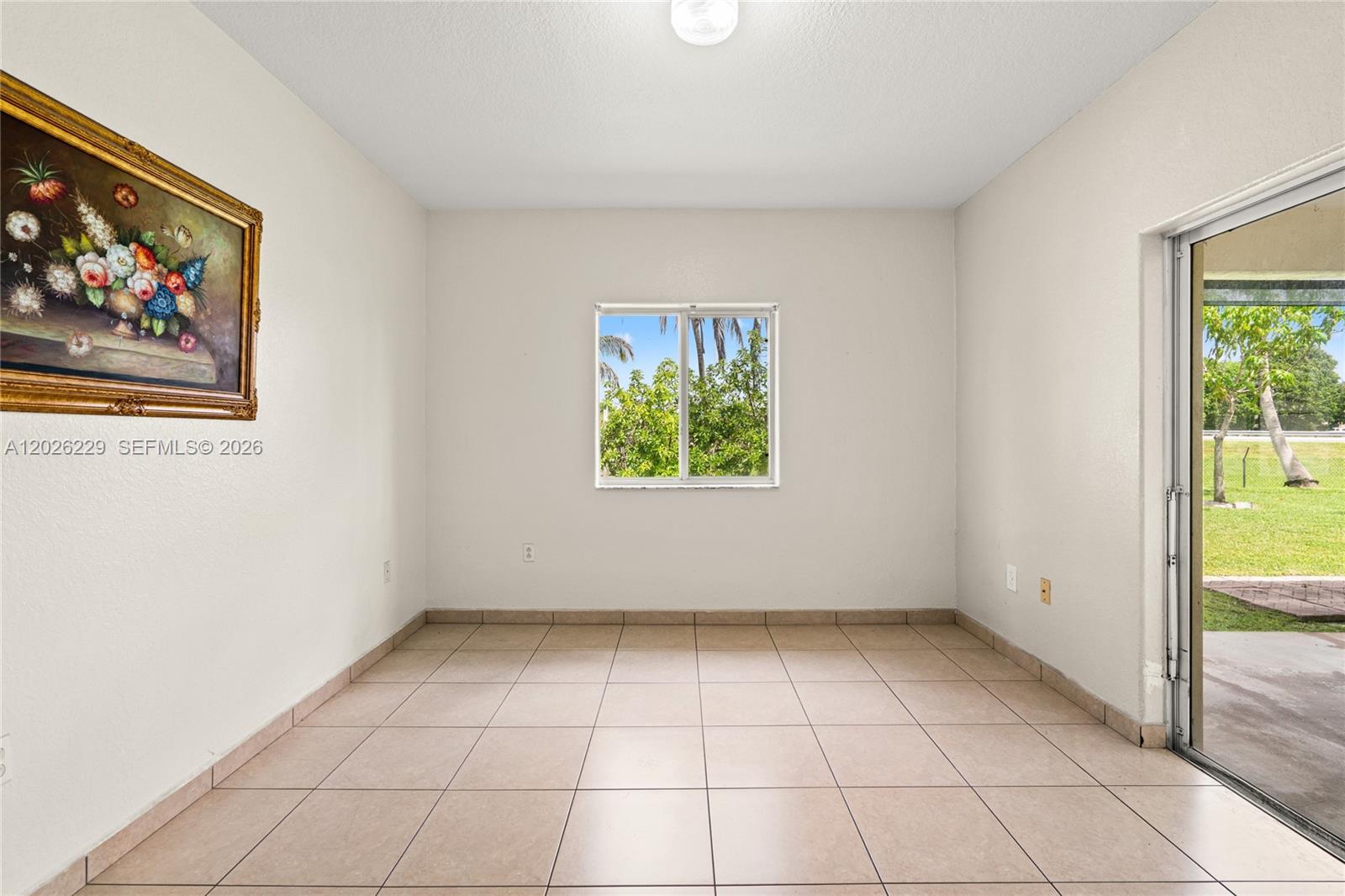 An empty room features beige tile flooring, a window, and a framed floral painting — 716 SE 12th Ter, Homestead FL 33033, HOMESTEAD, FL