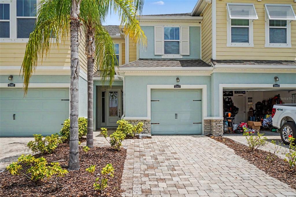 The exterior of a multi-unit townhouse features light blue garage doors, stone accents, and a palm tree — 12411 TRAILHEAD DRIVE, BRADENTON, FL
