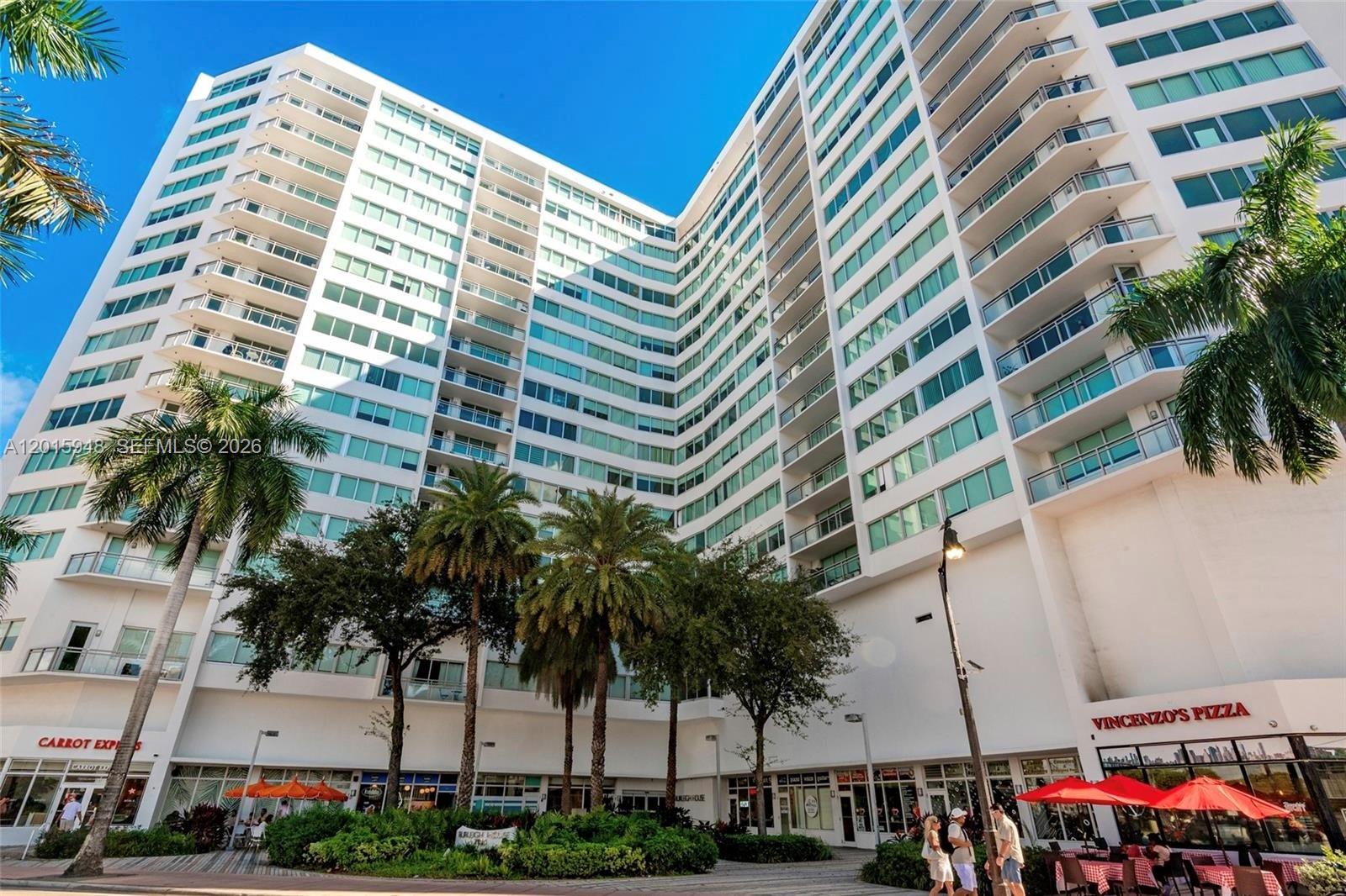 A low-angle view of a tall, white, multi-story apartment building with ground-floor retail shops — 7135 Collins Ave # 1214, Miami Beach FL 33141, MIAMI BEACH, FL