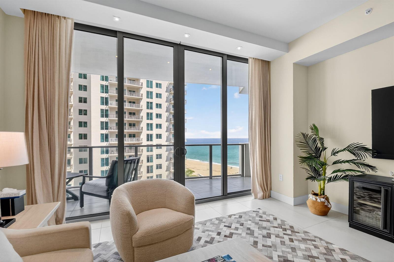 A living room with a beige armchair, patterned rug, and floor-to-ceiling windows overlooking the ocean — 3100 N Ocean Drive # H, Singer Island FL 33404, SINGER ISLAND, FL