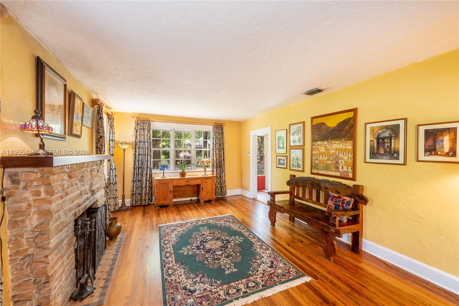 A living room features yellow walls, a brick fireplace, hardwood floors, and a wooden bench — 1256 Algeria Ave, Coral Gables FL 33134, CORAL GABLES, FL