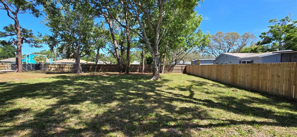 A grassy backyard features mature trees and a wooden privacy fence under a blue sky — 13296 98TH AVENUE N, SEMINOLE, FL