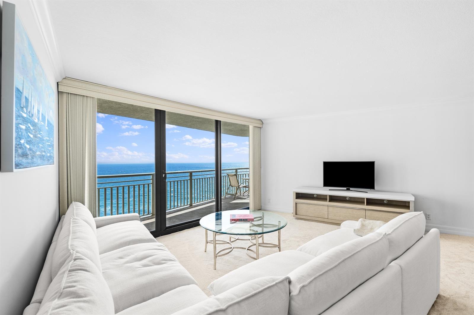 A living room features white sofas, a glass coffee table, and a balcony overlooking ocean — 3000 N Ocean Drive # 28e, Riviera Beach FL 33404, RIVIERA BEACH, FL