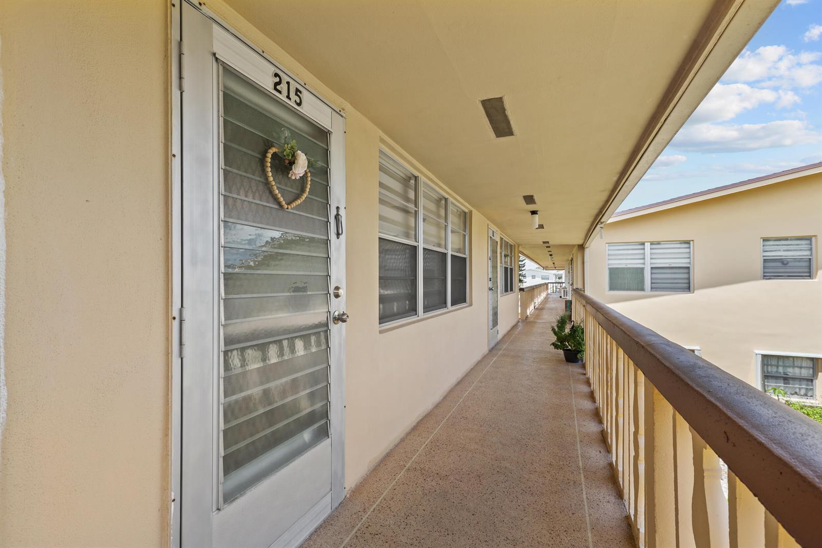 An outdoor apartment building walkway features a numbered door, windows, and a railing — 215 Sheffield I, West Palm Beach FL 33417, WEST PALM BEACH, FL