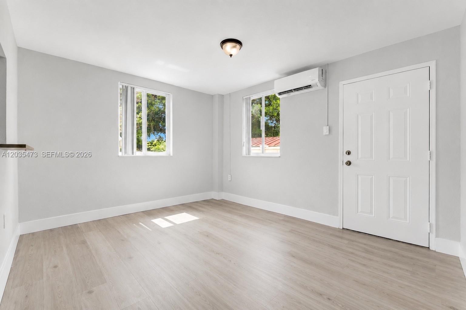 An empty room features light gray walls, wood-look flooring, two windows, and a wall-mounted air conditioner — 6647 SW 59th Pl # 101, South Miami FL 33143, SOUTH MIAMI, FL
