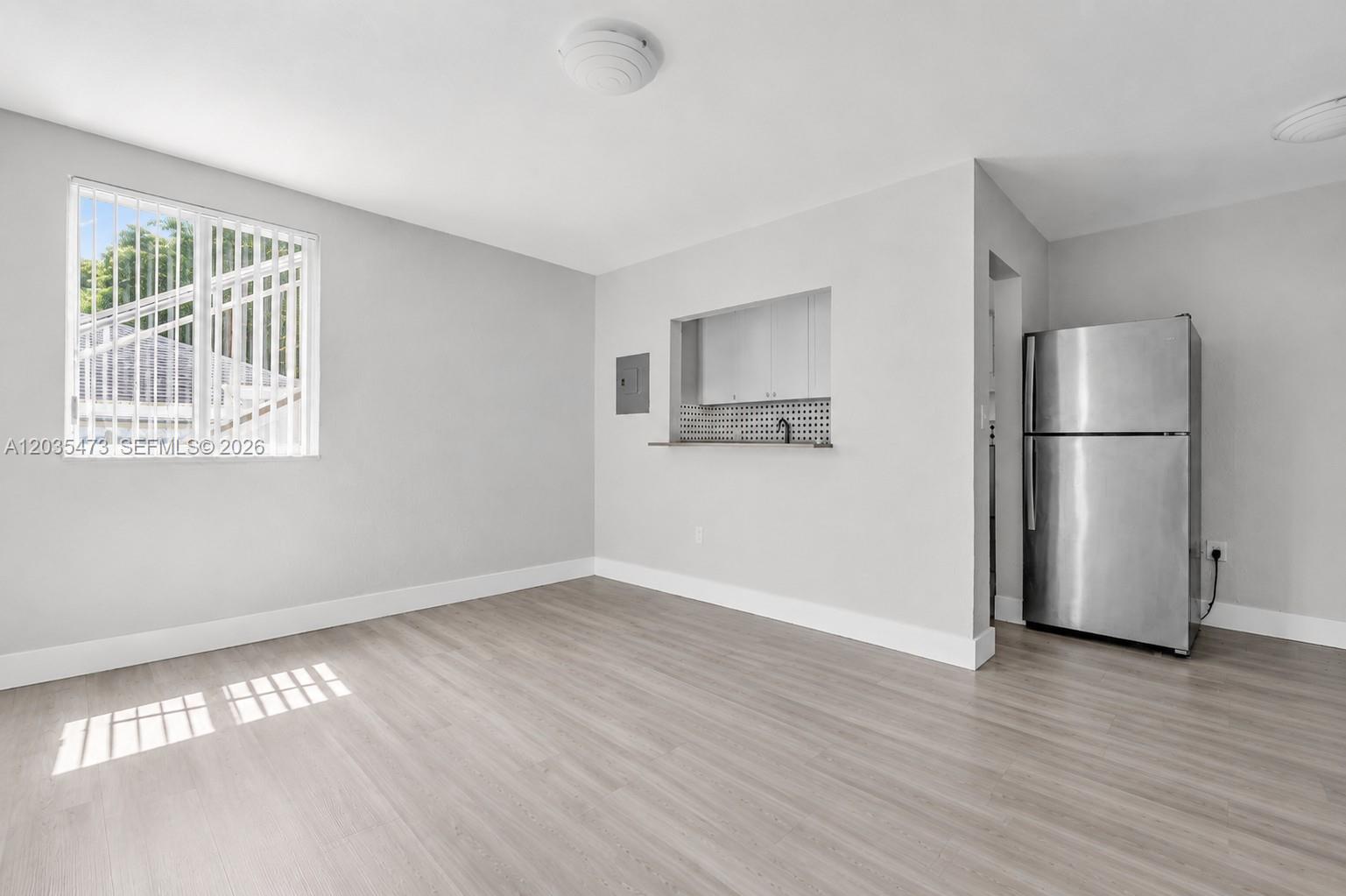 A room with light wood-look flooring, a window, a pass-through kitchen opening, and a refrigerator — 6647 SW 59th Pl # 101, South Miami FL 33143, SOUTH MIAMI, FL