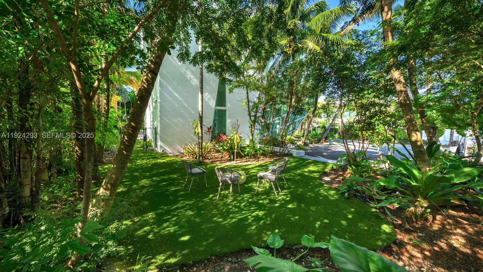 A grassy backyard features three outdoor chairs nestled among trees near a modern white building — 480 Ocean Blvd, Golden Beach FL 33160, GOLDEN BEACH, FL