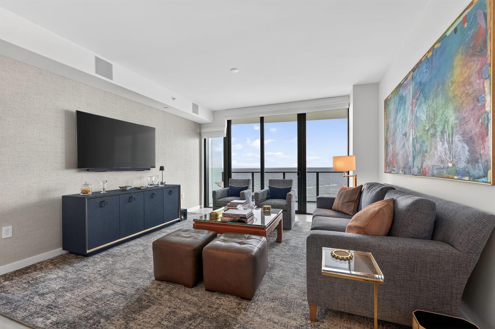 A living room with a grey sofa, blue media console, and floor-to-ceiling ocean view windows — 1116 N Ocean Boulevard # 1204, Pompano Beach FL 33062, POMPANO BEACH, FL