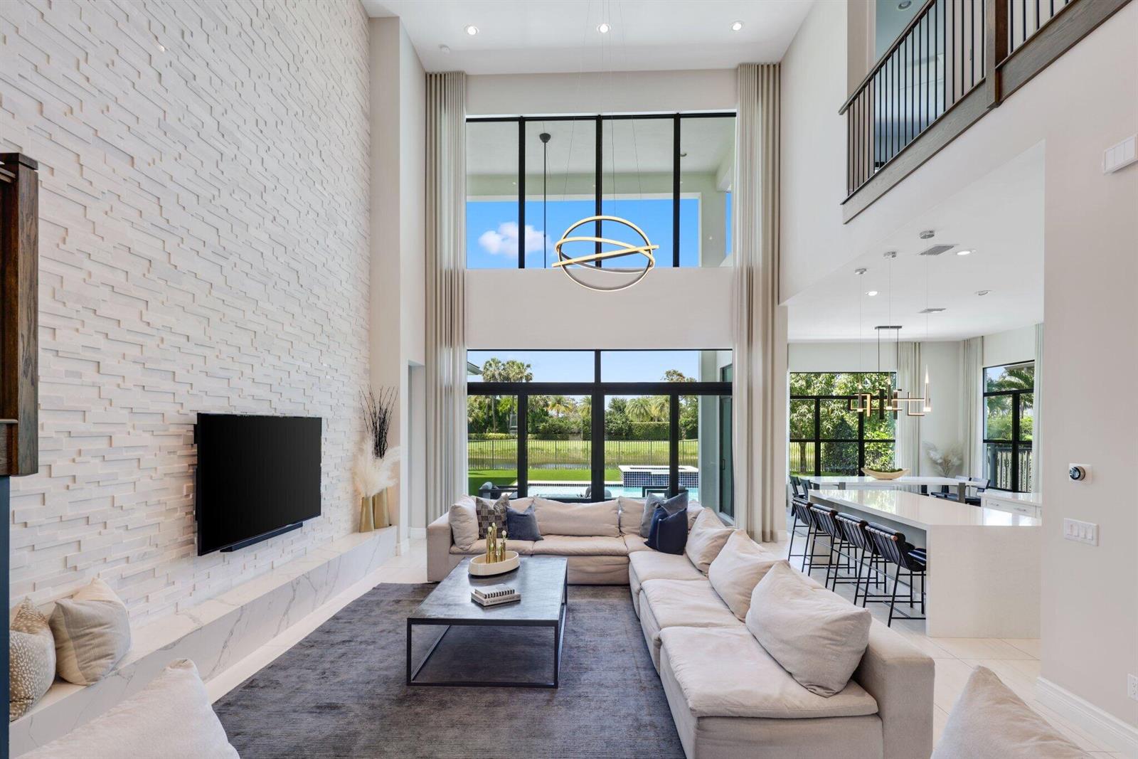 A double-height living room features a textured white stone wall, large windows, and a kitchen — 9275 Biaggio Road, Boca Raton FL 33496, BOCA RATON, FL