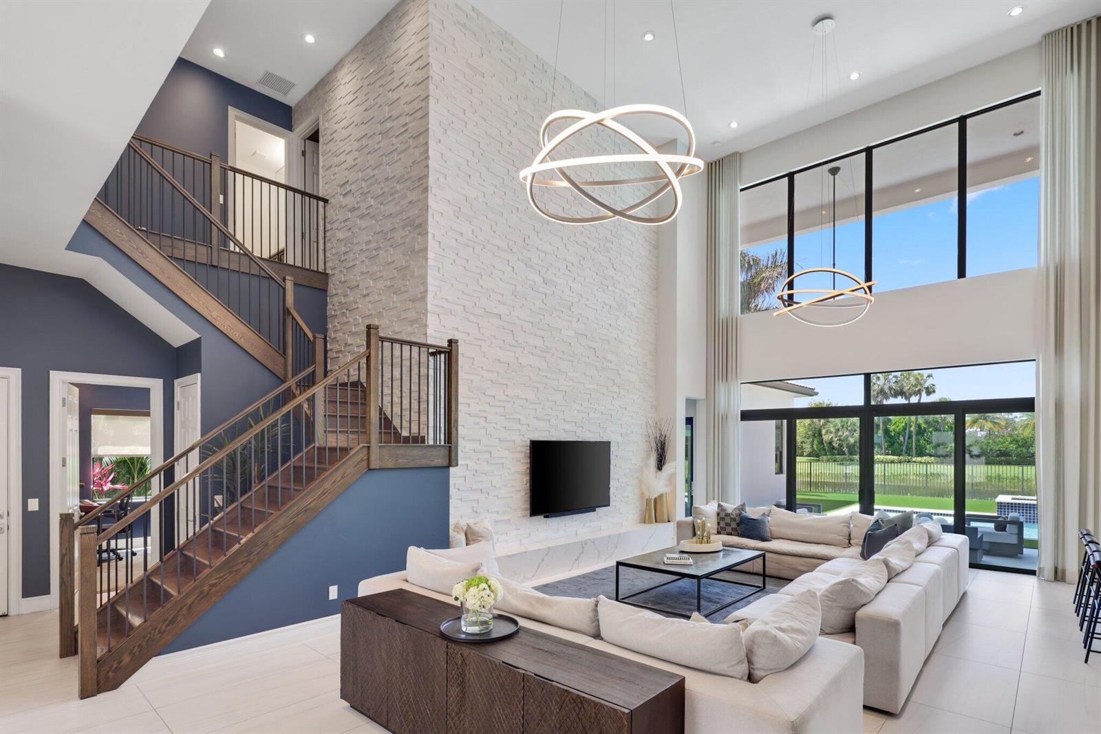 A modern living room features a two-story stone fireplace, a large sectional, and a staircase — 9275 Biaggio Road, Boca Raton FL 33496, BOCA RATON, FL