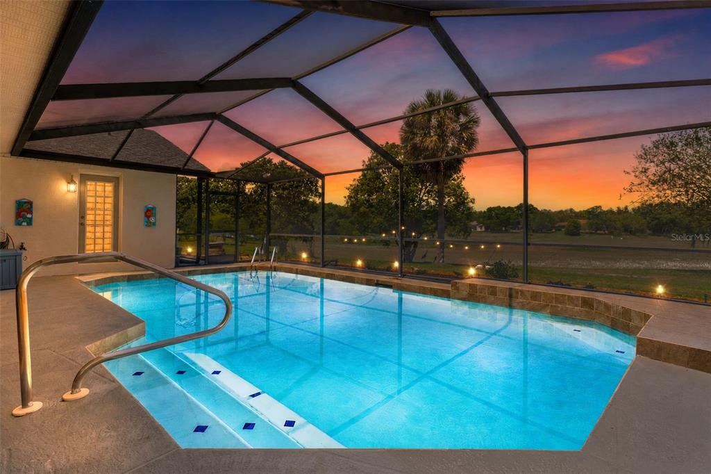 A screened-in swimming pool with steps overlooks a grassy field at sunset — 18652 AUTUMN LAKE BOULEVARD, HUDSON, FL