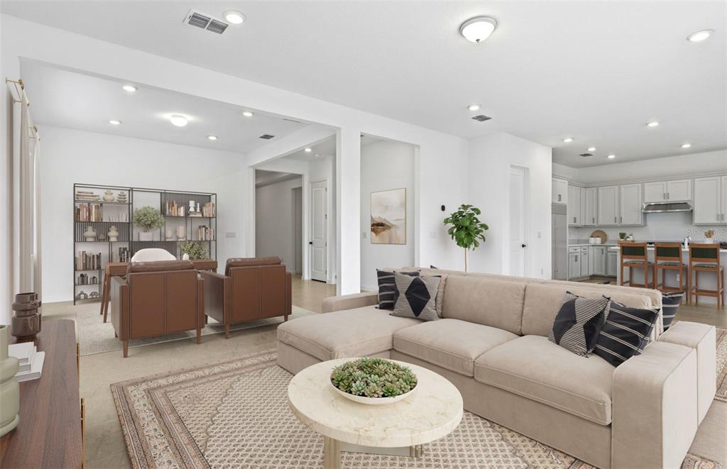A bright, open-concept living room features a beige sectional sofa, a round coffee table, and office furniture — 7046 DEPUE DRIVE, WESLEY CHAPEL, FL