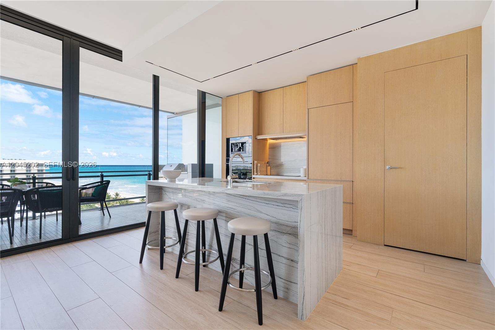 A modern kitchen features a marble waterfall island, light wood cabinetry, and a balcony view — 8701 Collins Ave # 1103, Miami Beach FL 33154, MIAMI BEACH, FL