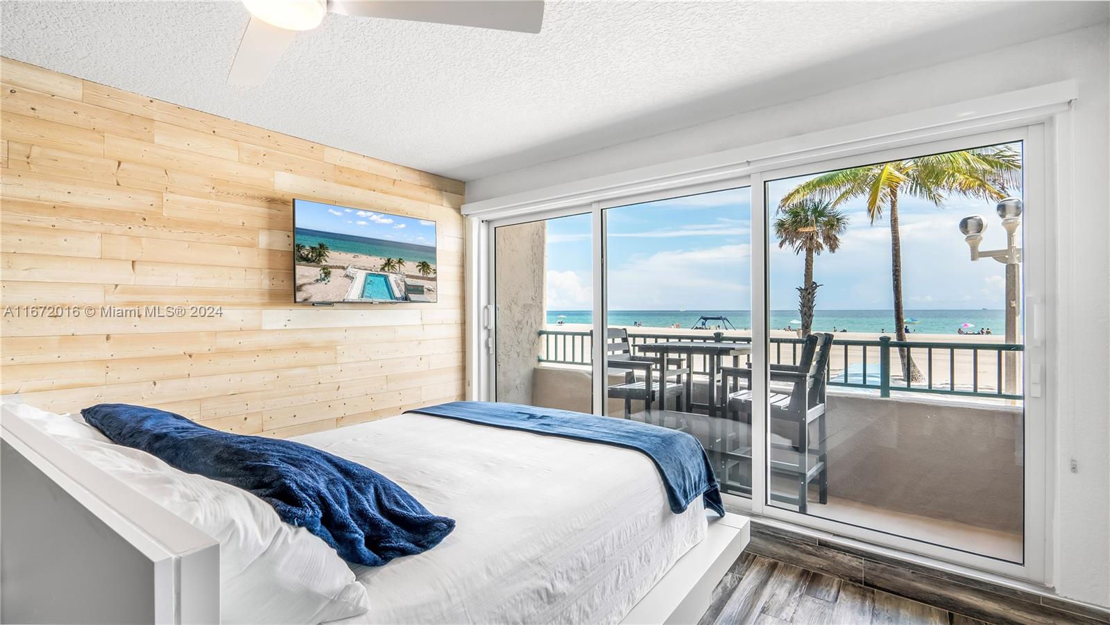 A bedroom features a wood-paneled wall, a bed with blue bedding, and a balcony overlooking the ocean — 201 Van Buren St # 103, Hollywood FL 33019, HOLLYWOOD, FL