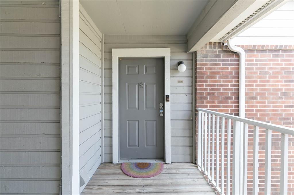 A gray front door with a rainbow doormat sits on a wooden balcony porch — 1400 GANDY BOULEVARD N UNIT 507, ST PETERSBURG, FL