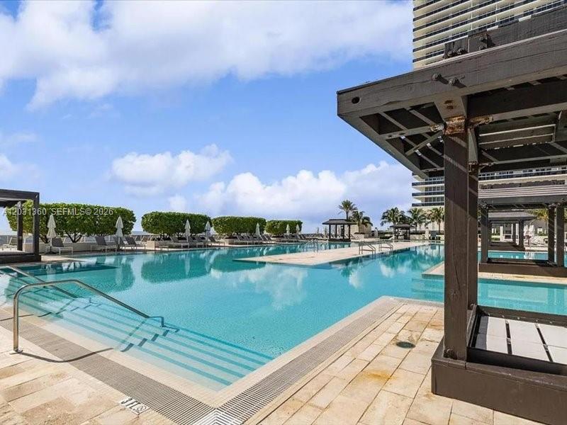 A large outdoor swimming pool features tiered steps and wooden cabanas on a sunny day — 1800 S Ocean Dr # 3101, Hallandale Beach FL 33009, HALLANDALE BEACH, FL