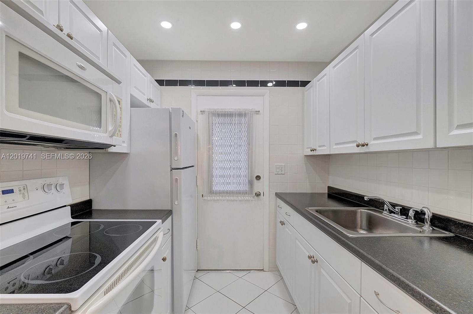 A galley kitchen features white cabinets, black countertops, a white refrigerator, and a glass-paneled door — 17001 NE 13th ave # 306, Miami FL 33162, MIAMI, FL