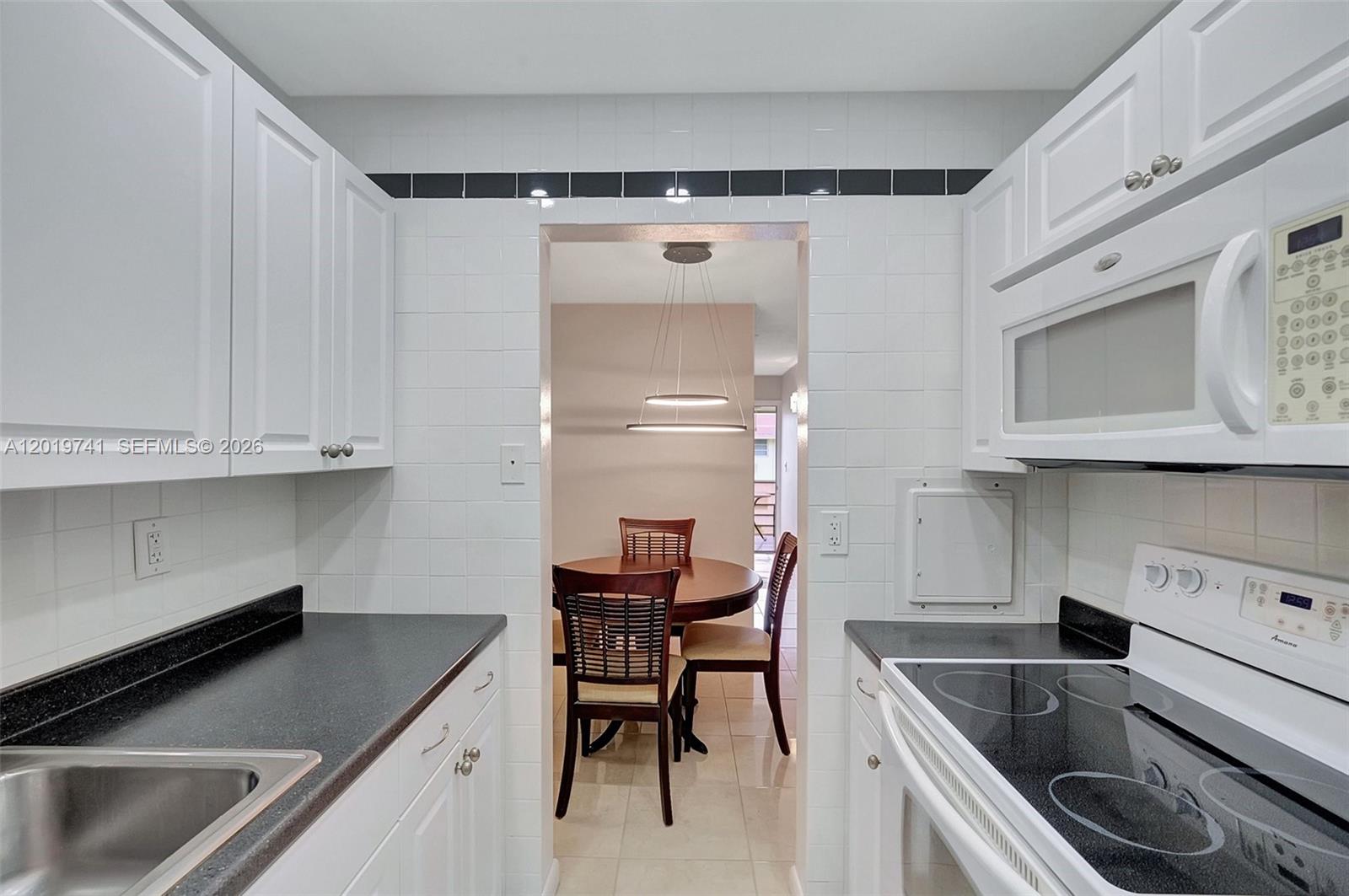 A galley kitchen with white cabinets and black countertops opens into a dining area — 17001 NE 13th ave # 306, Miami FL 33162, MIAMI, FL